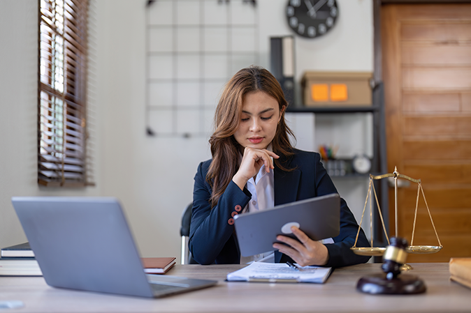 Young professional woman holding a tablet in front a laptop with the scales of justice and judge's gavel nearby