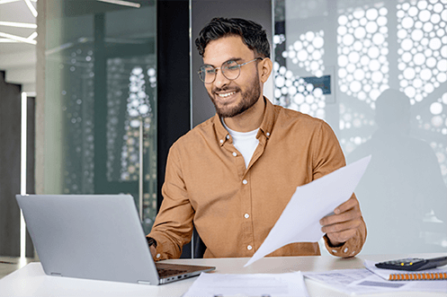 Young man wearing glasses holding a document while typing on a laptop