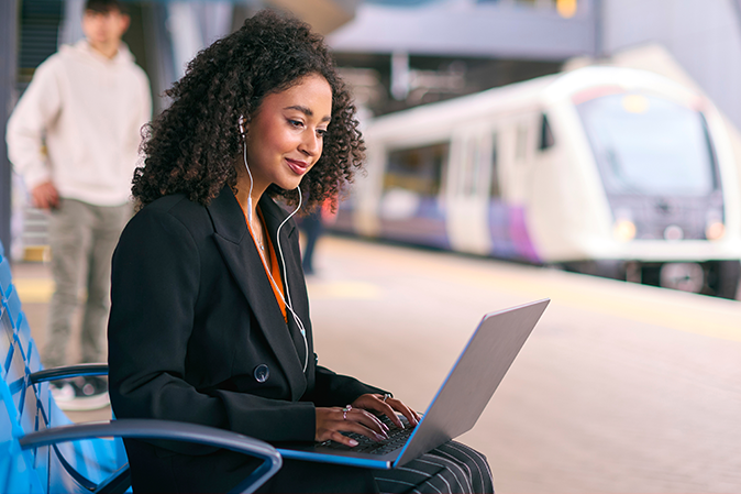 Business woman on a train platform working on laptop wearing earphones