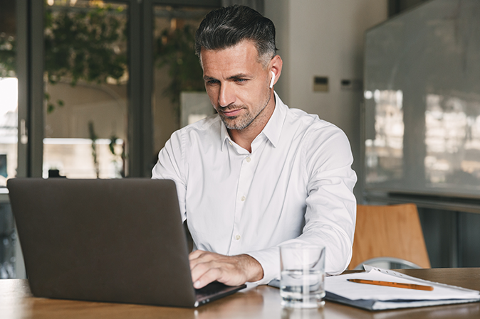 Photo of european businessman 30s wearing white shirt and wireless earphones sitting at table in office and working at laptop