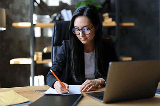 Young woman with glasses writing notes at a desk in front of a laptop