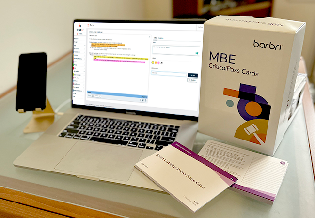 Laptop sitting on a desk with a mobile phone and the MBE CriticalPass Cards box set