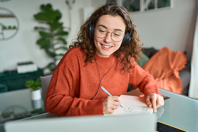 Young woman wearing headphones while watching a video on laptop and taking notes