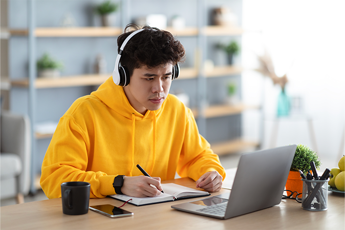 Young man wearing headphones while watching webinar on laptop and taking notes