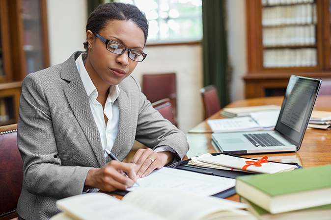 Young woman dressed professionally writing notes at a desk in front of a laptop
